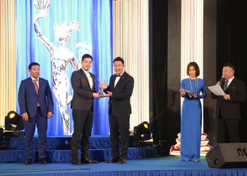 Шагнал гардуулах ёслол дээр албан ёсны хувцастай тайзан дээрх хүмүүс; бусад нь харж байхад өөр хүнд цом гардуулж буй хүн. - Business.MN