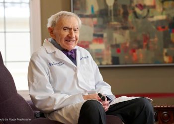 Лабораторийн цагаан цув өмссөн өндөр настан сандал дээр инээмсэглэн сууж байна. Тэд зүүн гартаа хар юм шиг харагддаг зүйлийг барьдаг. Цаана нь ханан дээр хийсэн хийсвэр зураг, байгалийн гэрэл оруулах том цонх байдаг нь энтрепренерт тохирсон урам зориг өгөх уур амьсгалыг бүрдүүлдэг. - Business.MN