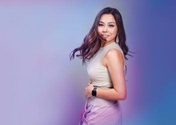 Урт, долгион үстэй эмэгтэй градиент хөх, нил ягаан өнгийн дэвсгэр дээр зогсож байна. Тэрээр ханцуйгүй шаргал цамц, өндөр бэлхүүстэй ягаан өмд өмсөж, мөрөн дээгүүрээ хараад инээмсэглэдэг. Түүний ухаалаг цаг, том ээмэг нь энтрепренерийн сүнсийг шингээсэн боловсронгуй байдлыг нэмж өгдөг. - Business.MN