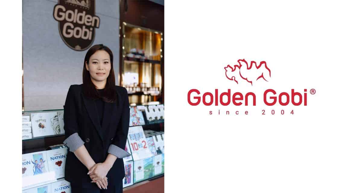 Урт хар үстэй, хар пиджак өмссөн эмэгтэй номын тавиур болон "Алтан говь" гэсэн самбарын өмнө зогсож байна. Энтрепренерийн хувьд тэрээр Алтан говийн бизнесийг бахархалтайгаар төлөөлдөг. Зургийн баруун талд "Алтан говь 2004 оноос хойш" гэсэн бичвэр, хоёр тэмээний тойм бүхий Алтан говийн лого дүрслэгдсэн байна. - Business.MN