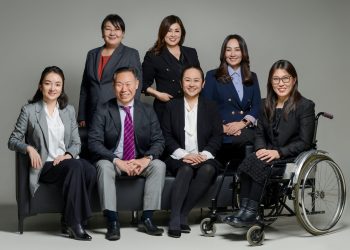 Дөрвөн эмэгтэй зогсож, гурван хүн сууж буй бизнесийн долоон мэргэжилтнүүдийн нэг нь нил ягаан өнгийн зангиа зүүсэн эрэгтэй, нөгөө нь тэргэнцэртэй энтрепренер бөгөөд бүгд камерын өмнө харан, саармаг саарал дэвсгэртэй студийн орчинд инээмсэглэж байна. - Business.MN