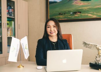 Оффис дээр зөөврийн компьютертэй ширээний ард сууж буй эмэгтэй. Тэр хар хүрэм өмсөж, камер руу инээмсэглэн, хуваарийнхаа хоосон орон зайг хэрхэн дүүргэх талаар бодож байгаа байх. Ширээг хоёр туг чимж, арын ханыг ландшафтын зураг чимдэг. - Business.MN