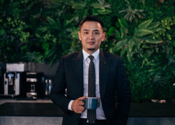 Костюм өмссөн эр аяга бариад лангуу налан мэргэжлийн бизнесийн аура гаргаж байна. Түүний ард янз бүрийн ургамлаар чимэглэсэн өтгөн ногоон хана байх бөгөөд лангуун дээр RE/MAX загвартай кофены машинууд эгнэн зогсоно. - Business.MN