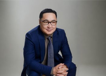 Цэнхэр костюмтай, зангиа зүүсэн хүн гараа атгасан байдалтай урагш бага зэрэг бөхийж сууна. Тэд нүдний шил зүүж, энгийн, цайвар саарал дэвсгэр дээр нарийн инээмсэглэдэг бөгөөд энэ нь бизнесийн жинхэнэ амжилтыг эд баялагаар бус, хэдэн хүний амьдралыг өөрчилснөөр хэмжигддэг гэсэн санааг илэрхийлдэг. - Business.MN