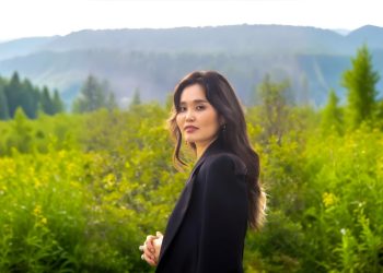 Үүлэрхэг тэнгэрийн дор уулс, модоор хүрээлэгдсэн шар зэрлэг цэцэгсийн өтгөн ногоон талбайд урт үстэй эмэгтэй зогсож байна. Хар хүрэм өмссөн тэрээр дотоод хөтөчөөр удирдуулсан мэт байгалийн амгалан тайван байдлыг шингээсэн камер руу ширтэнэ. - Business.MN