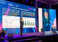 Монголын хөдөлмөрийн зах зээлийн чуулга уулзалт 2024 дээр илтгэгч тайзан дээр зогсож асуудал, шийдлийг том дэлгэц дээр харуулсан графикаар танилцуулж байна. Тэд микрофон руу ярьж байгаа бол өөр дэлгэц нь шууд видео бичлэгийг харуулж байна. Тайзны гэрэлтүүлэг, төрөл бүрийн баннерууд нь сэтгэл татам уур амьсгалыг бүрдүүлдэг. - Business.MN