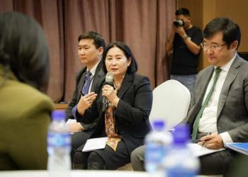A woman speaks into a microphone while sitting in a discussion group. Two men sit beside her, and other participants and a photographer are in the background. Bottles of water are visible on a table in the foreground.